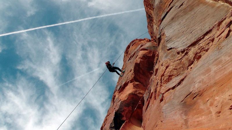 A rock climber rappelling down a tall, reddish rock face against a backdrop of a bright blue sky with wispy clouds. The climber is secured with ropes and appears to be descending carefully. The rock surface shows various textures and crevices, highlighting the rugged terrain.