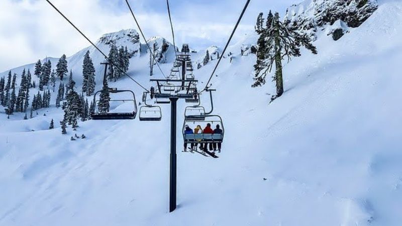 A ski lift carrying people over a snowy mountain terrain, with rocky peaks and scattered evergreen trees in the background.