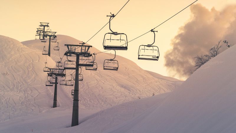 A row of chairlifts suspended over snow-covered slopes at sunset, with tracks from previous skiers visible in the background.