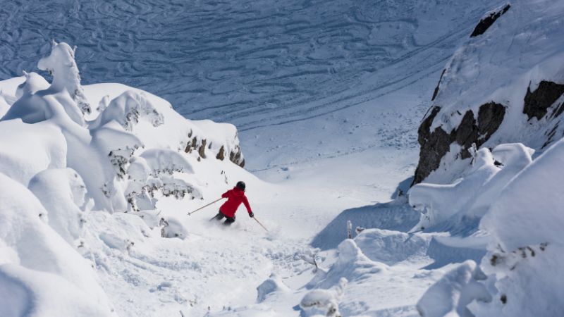 A skier wearing a red jacket descends a steep, narrow chute surrounded by snow-covered rock formations in a remote mountain setting.